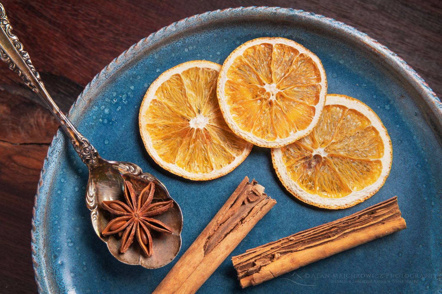 Spices and dried orange slices on blue stoneware plate, with cinnamon sticks, and star anise resting on an antique spoon #79063