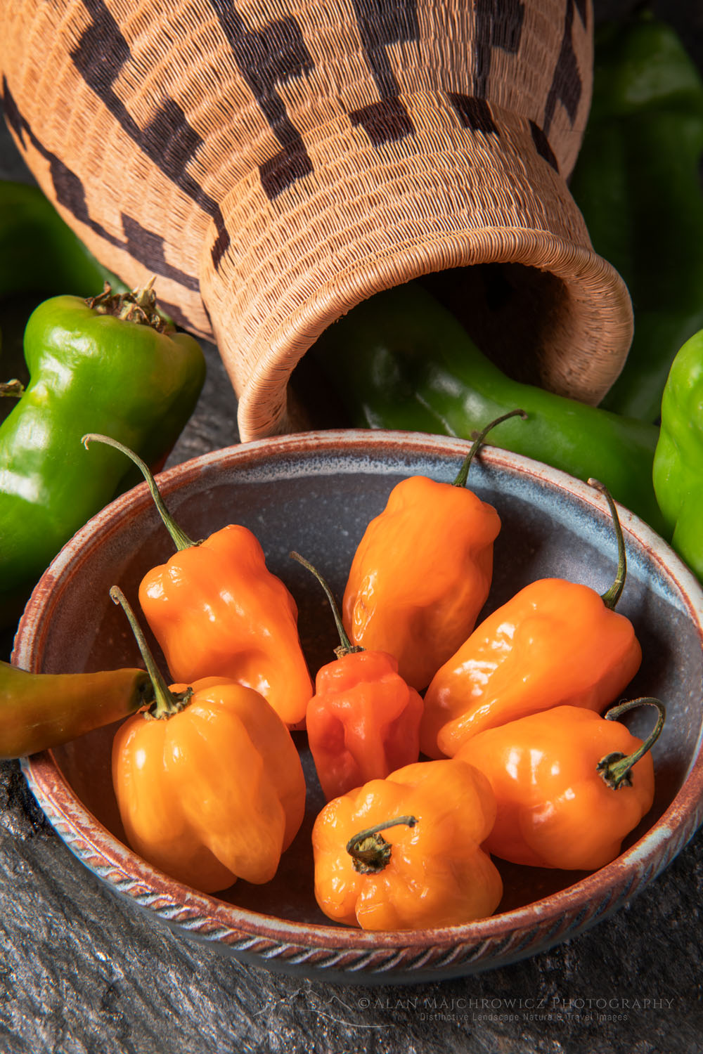 Arrangement of Habanero and Hatch chile peppers with Panamanian Wounaan basket and stoneware bowl on stone backdrop #81175